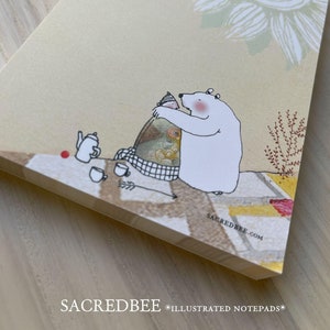 May include: A notepad with a whimsical illustration of a white polar bear hugging a colorful object. The notepad has a light beige background with a large white sunflower design and the text "SACREDBEE.COM".