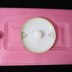 May include: A pink painted cabinet door with a white porcelain knob in the center. The door has two white porcelain knobs on either side of the center knob. A white porcelain plate with a gold rim and floral design is centered on the door.