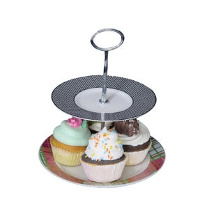 Op de afbeelding: Een tweelaagse taartstandaard met een zilveren handvat en zwart-wit geblokte borden. De standaard is gevuld met vier cupcakes, een met groene frosting en een roze bloem, een met witte frosting en kleurrijke sprinkles, een met witte frosting en een chocolade reep en een met chocolade frosting.