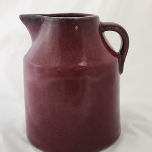 May include: A burgundy ceramic pitcher with a handle and a spout. The pitcher has a cylindrical body and a dark rim. The handle is a loop shape. The pitcher is likely used for serving beverages.