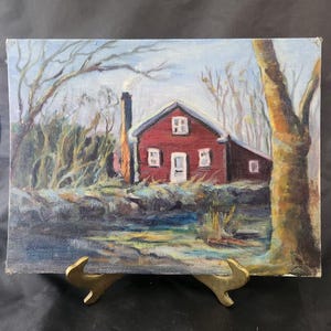 May include: Oil painting of a red house with a black chimney, set in a winter landscape. Bare trees frame the house, with a stream in the foreground. The painting is displayed on a gold-colored easel.