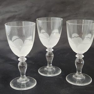 May include: Three clear wine glasses with a frosted floral design on the upper portion of the bowl. The glasses have a rounded base, a decorative stem, and a clear, smooth finish. The background is a dark, solid color.