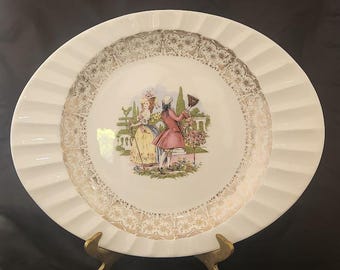 May include: A white decorative plate with a ruffled edge and a gold filigree border. The center features a colorful illustration of two figures in period clothing in a garden setting. The plate is displayed on a gold stand.