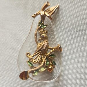 May include: A vintage pear-shaped brooch with a clear, glass-like body. A gold-colored bird perches on a branch with green leaves and small red accents. The brooch has a gold-colored stem and leaf detail.