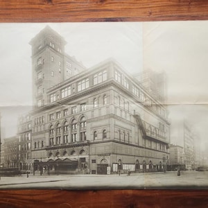 May include: Vintage black and white photograph of a large building with multiple stories, arched windows, and a tower. The image is slightly faded, suggesting age. The building is on a city street.