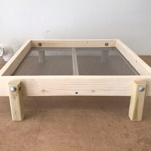 May include: A wooden sifting tray with a fine mesh screen. The frame is made of light-colored wood and is supported by four short legs. A white PVC pipe fitting is visible in the background.
