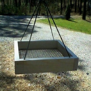 May include: A square, wooden fire pit with a metal grate base, suspended by black metal cables. The fire pit is light brown and appears to be outdoors, with a gravel surface and greenery in the background.