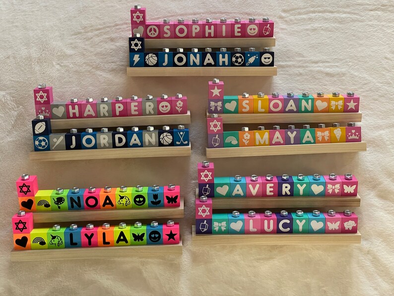 Puede incluir: Un conjunto de ocho menor&aacute;s de madera de colores, cada una con un nombre diferente escrito en la parte delantera. Los nombres son: Sophie, Jonah, Harper, Jordan, Sloan, Maya, Avery y Lucy. Cada menor&aacute; tiene una variedad de peque&ntilde;os s&iacute;mbolos en los bloques, incluyendo estrellas, corazones, arco&iacute;ris y mariposas.