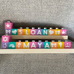Puede incluir: Bloques de madera que deletrean los nombres "Sloan" y "Maya" con varios dise&ntilde;os y s&iacute;mbolos coloridos en cada bloque.