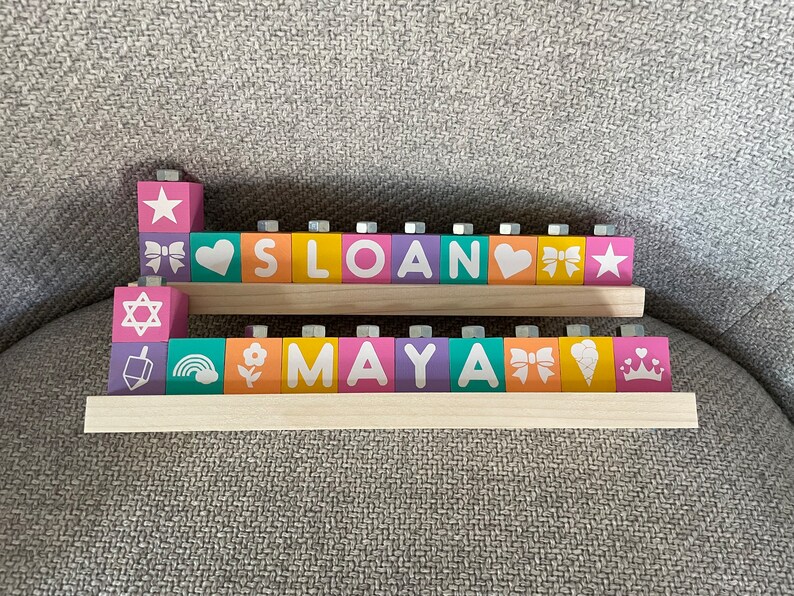 Puede incluir: Bloques de madera que deletrean los nombres "Sloan" y "Maya" con varios dise&ntilde;os y s&iacute;mbolos coloridos en cada bloque.