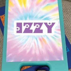 May include: A teal blue clipboard with a tie-dye design and a purple sticker with the word "IZZY" in white letters. The sticker has small white flowers on either side of the word.