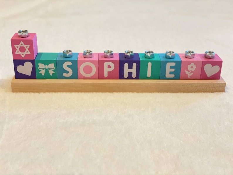 Puede incluir: Una menor&aacute; de madera con bloques coloridos que deletrean el nombre "SOPHIE". Los bloques tienen diferentes dise&ntilde;os, incluyendo una estrella de David, un coraz&oacute;n, un lazo, una flor y un coraz&oacute;n.