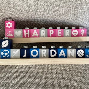 Puede incluir: Dos filas de bloques de madera que deletrean los nombres "HARPER" y "JORDAN". Los bloques est&aacute;n pintados de rosa, gris y azul con letras y s&iacute;mbolos blancos. La fila superior tiene una estrella de David, un coraz&oacute;n, una corona y una cara sonriente. La fila inferior tiene un bal&oacute;n de f&uacute;tbol, un bal&oacute;n de f&uacute;tbol, un bate de b&eacute;isbol y un bal&oacute;n de baloncesto.