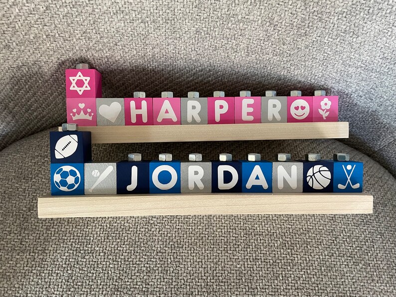 Puede incluir: Dos filas de bloques de madera que deletrean los nombres "HARPER" y "JORDAN". Los bloques est&aacute;n pintados de rosa, gris y azul con letras y s&iacute;mbolos blancos. La fila superior tiene una estrella de David, un coraz&oacute;n, una corona y una cara sonriente. La fila inferior tiene un bal&oacute;n de f&uacute;tbol, un bal&oacute;n de f&uacute;tbol, un bate de b&eacute;isbol y un bal&oacute;n de baloncesto.