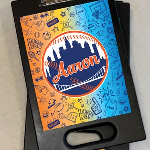 May include: Black clipboard with a colorful graphic design. The design features a baseball theme with the name "Aaron" in a baseball-shaped design. The background is split between orange and blue with sports-related illustrations.