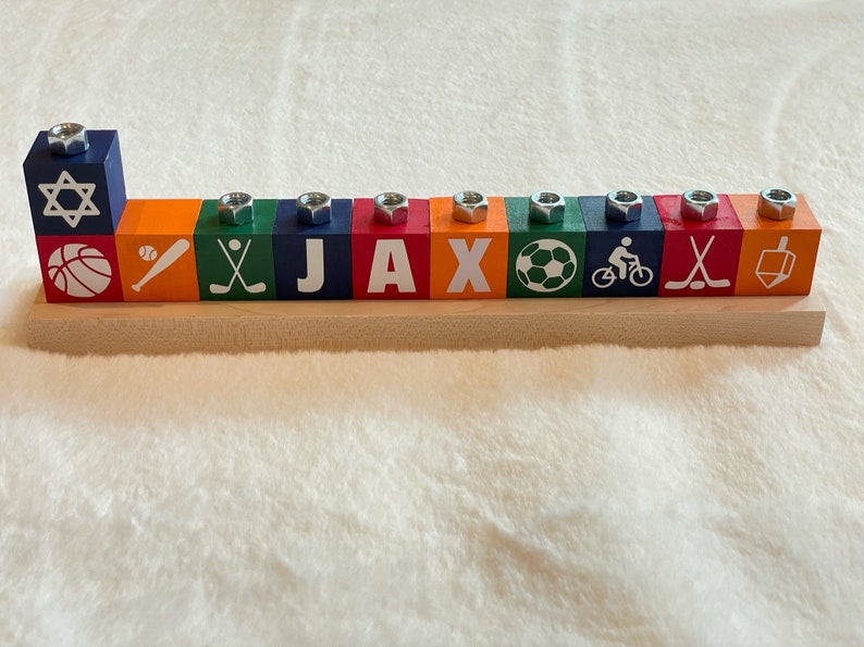 Puede incluir: Una menor&aacute; de madera con nueve bloques azules, naranjas y verdes. Cada bloque tiene una imagen diferente con tem&aacute;tica deportiva y una tuerca plateada en la parte superior. Los bloques deletrean el nombre "JAX".