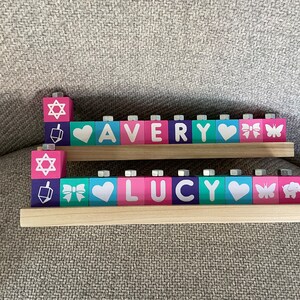 Puede incluir: Una menor&aacute; de madera con dos filas de bloques que deletrean los nombres "AVERY" y "LUCY". Cada bloque tiene un color diferente y un dise&ntilde;o, que incluye una estrella de David, un coraz&oacute;n, un lazo, una mariposa y un elefante.