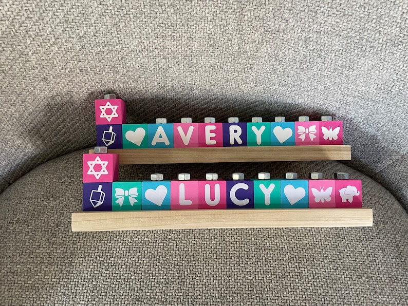 Puede incluir: Una menor&aacute; de madera con dos filas de bloques que deletrean los nombres "AVERY" y "LUCY". Cada bloque tiene un color diferente y un dise&ntilde;o, que incluye una estrella de David, un coraz&oacute;n, un lazo, una mariposa y un elefante.