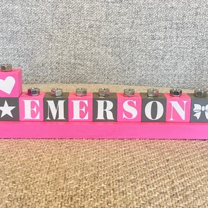 Puede incluir: Bloques de madera rosa con el nombre "EMERSON" escrito en letras blancas. Los bloques tienen un fondo gris y cada bloque tiene un dise&ntilde;o diferente en la parte superior, incluyendo un coraz&oacute;n, una estrella, un lazo y una tuerca.