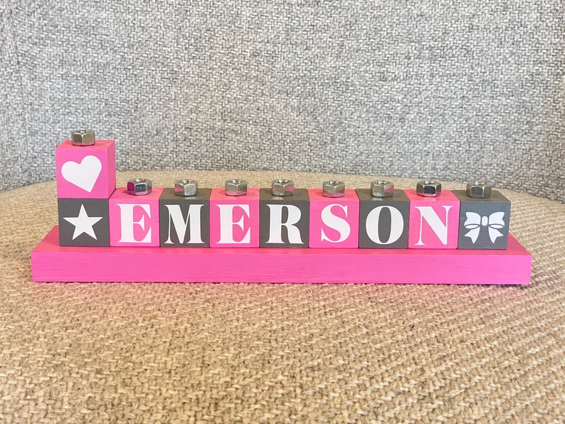 Puede incluir: Bloques de madera rosa con el nombre "EMERSON" escrito en letras blancas. Los bloques tienen un fondo gris y cada bloque tiene un dise&ntilde;o diferente en la parte superior, incluyendo un coraz&oacute;n, una estrella, un lazo y una tuerca.