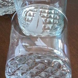 May include: Two clear glass tumblers with a textured bottom and etched sailboat design. The bottom tumbler has the text "True North Blanc Nord" etched on the side.