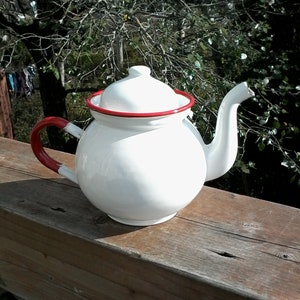 May include: White enamel teapot with a red rim and handle. The teapot has a round body and a spout on the side.