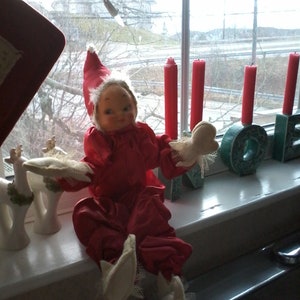 Peut inclure: Une poupée d'elfe rouge et blanche avec une bordure en dentelle blanche est assise sur un rebord de fenêtre. La poupée a un visage blanc avec des traits peints et porte une robe rouge avec une bordure en dentelle blanche. La poupée tient un objet blanc et rouge dans sa main. Il y a des bougies rouges en arrière-plan.