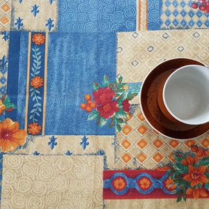 May include: A white coffee cup with a brown saucer on a colorful patterned tablecloth. The tablecloth has a patchwork design with floral and geometric patterns in shades of blue, orange, red, and beige.