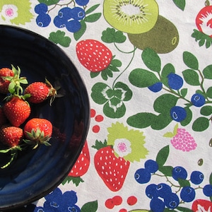 May include: A blue ceramic plate with a white rim holds a group of fresh red strawberries. The plate is on a white tablecloth with a colorful fruit pattern.