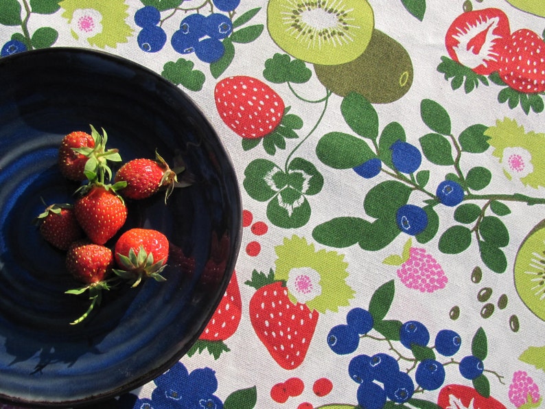 May include: A blue ceramic plate with a white rim holds a group of fresh red strawberries. The plate is on a white tablecloth with a colorful fruit pattern.