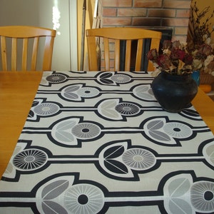 May include: A black and white geometric patterned table runner with a floral design. The runner is on a wooden table with a vase of dried flowers.