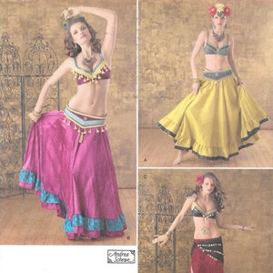 May include: Three women wearing different styles of belly dancing costumes. The costumes include a bra top, a belt, and a long skirt. The skirts are in different colors, including pink, yellow, and red. The women are posing in front of a decorative screen.