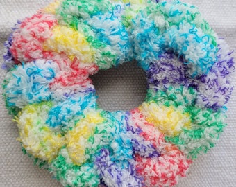May include: A circular hair scrunchie made of fuzzy, multi-colored yarn. The texture is soft and plush, featuring a vibrant mix of white, yellow, pink, blue, green, and purple strands woven together in a dense, curly, and voluminous pattern.