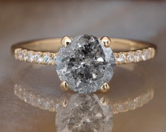 Puede incluir: Un anillo de compromiso de oro con un gran diamante gris redondo engastado en pavé. El anillo está sobre una superficie de mármol blanco.