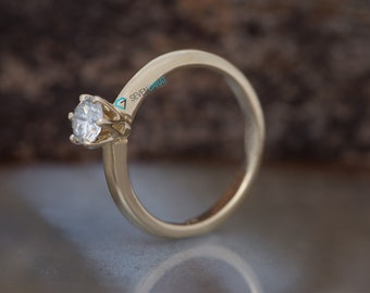 May include: A gold-colored engagement ring with a single, clear diamond. The ring has a classic design with a round diamond set in a four-prong setting. The band is smooth and polished. The text "SEVEN CARAT" is visible on the ring.