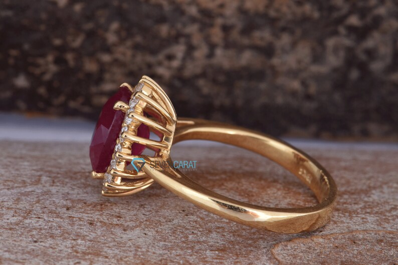 Puede incluir: Un anillo de oro con un gran rub&iacute; de forma ovalada rodeado por un halo de diamantes m&aacute;s peque&ntilde;os. El anillo est&aacute; engastado en un estilo cl&aacute;sico con una banda lisa.