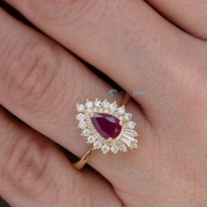 Puede incluir: Un anillo de oro con una piedra de rubí en forma de pera rodeada de diamantes más pequeños. El anillo está engastado en un diseño delicado de inspiración floral.