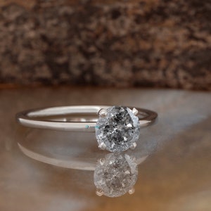 Puede incluir: Un anillo de compromiso de plata con un diamante redondo gris.