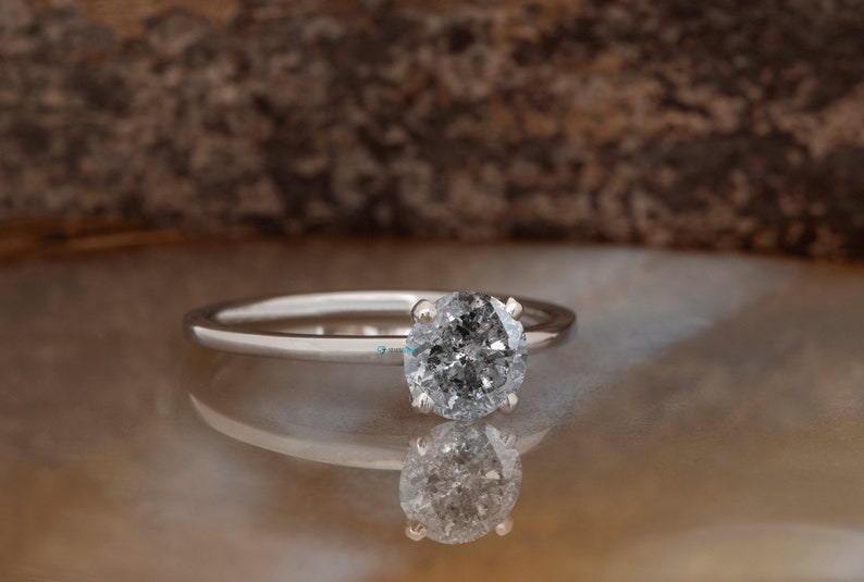 Puede incluir: Un anillo de compromiso de plata con un diamante redondo gris.