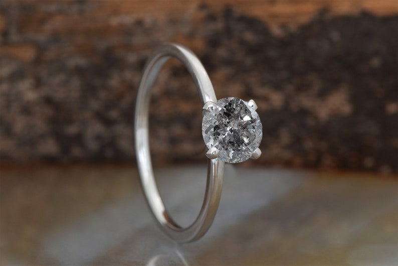 Puede incluir: Un anillo de compromiso de plata con un diamante redondo gris.