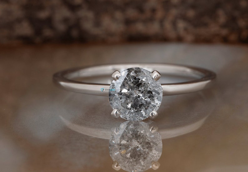 Puede incluir: Un anillo de compromiso de plata con un diamante redondo gris.