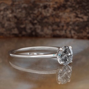 Puede incluir: Un anillo de compromiso de plata con un diamante redondo gris.