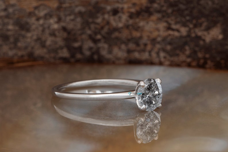 Puede incluir: Un anillo de compromiso de plata con un diamante redondo gris.