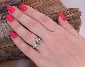 Puede incluir: Un anillo de compromiso de diamante gris con una banda pavé engastada en oro amarillo. El anillo está en un dedo con esmalte de uñas rojo.