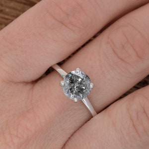 Puede incluir: Un anillo de compromiso de plata con un gran diamante redondo gris.