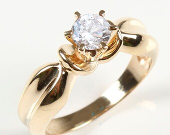 Op de afbeelding: Een gouden ring met een enkele, ronde, heldere edelsteen gezet in een knoopvormig ontwerp.