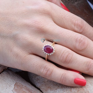 Puede incluir: Un anillo de oro con una gran piedra de rub&iacute; ovalada rodeada de diamantes m&aacute;s peque&ntilde;os. El anillo est&aacute; en un dedo con esmalte de u&ntilde;as rojo.