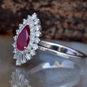 Puede incluir: Un anillo de rubí en forma de pera engastado en una banda de plata con un halo de diamantes más pequeños. El anillo está sobre una superficie blanca.