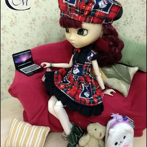 May include: A doll wearing a red and black tartan dress and a red and black tartan hat with a white bird design. The doll is sitting on a red sofa with a laptop computer on the sofa beside her. The doll is holding a small teddy bear and a white plush bag with a pink bow.