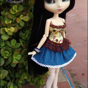 Può includere: Una bambola con lunghi capelli neri che indossa un cappello all'uncinetto bianco, un corpetto a stampa floreale, una gonna di jeans con bordo in pizzo e stivali bianchi.